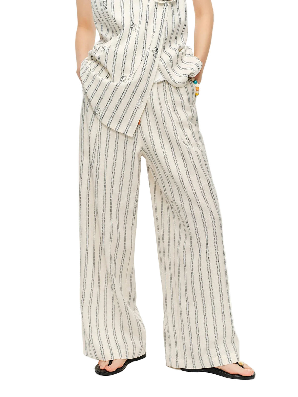A model wearing a sleeveless cream-colored jumpsuit with thin black vertical stripes and wide-leg pants, paired with black flip-flops. The Dushu brand logo is visible in the top-left corner against a plain white background.
