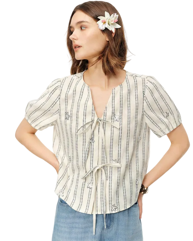 A woman wearing a short-sleeve striped blouse with a tie-front detail and drawstring, paired with light blue jeans. A white flower is tucked into her hair.